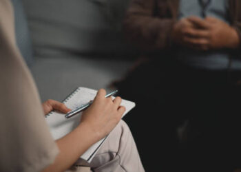 Portrait of Asian therapists writing down notes during therapy with male patients, taking notes in their office setting, addressing themes of depression and mental health support.